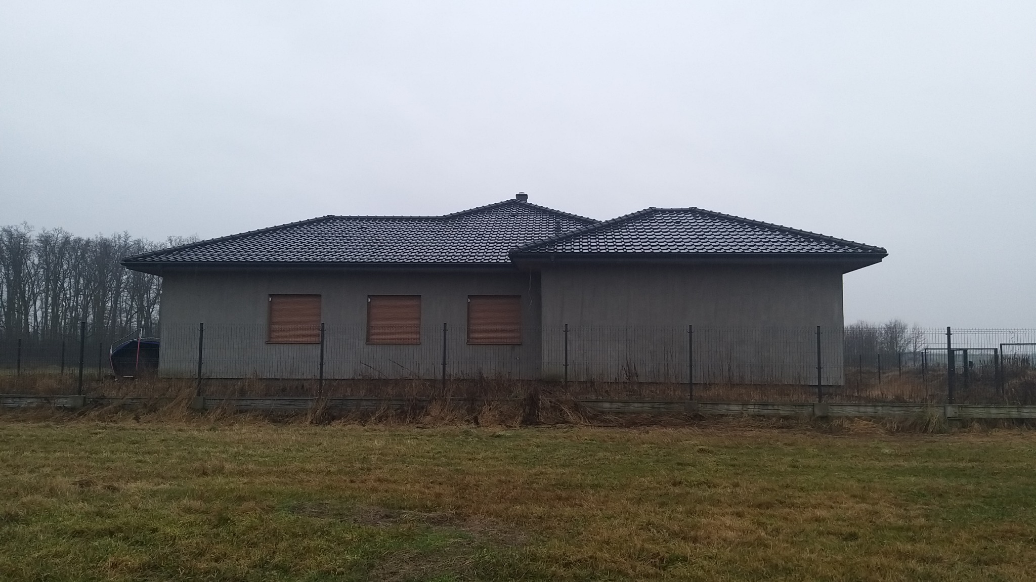 Surowy dom murowany z ciemną dachówką i trzema oknami z roletami, otoczony ogrodzeniem z siatki, widoczny na tle pochmurnego nieba i jesiennego krajobrazu.