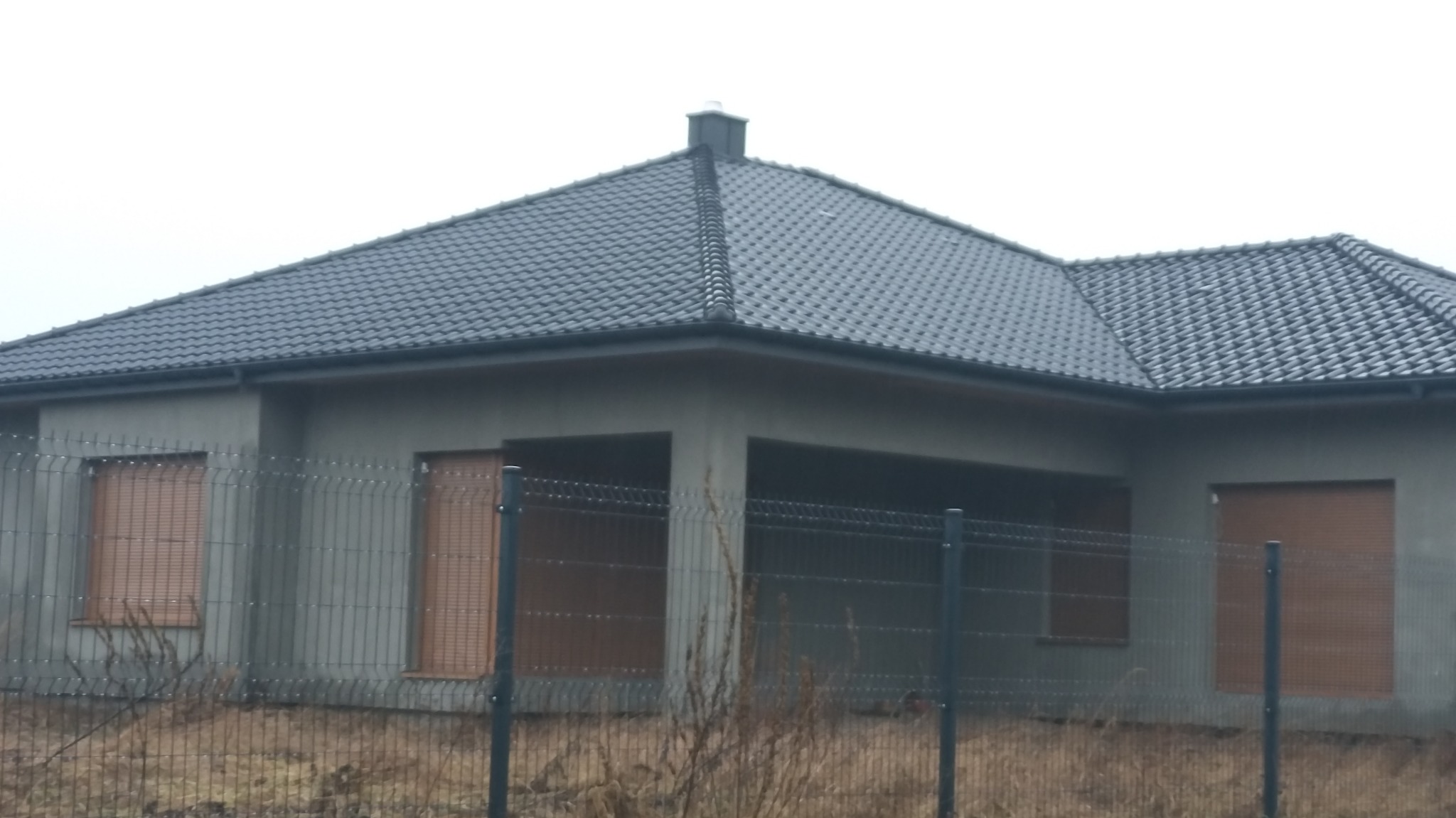 Surowy stan domu jednorodzinnego z ciemną dachówką, widoczny przez metalowe ogrodzenie tymczasowe, okna z roletami zewnętrznymi.