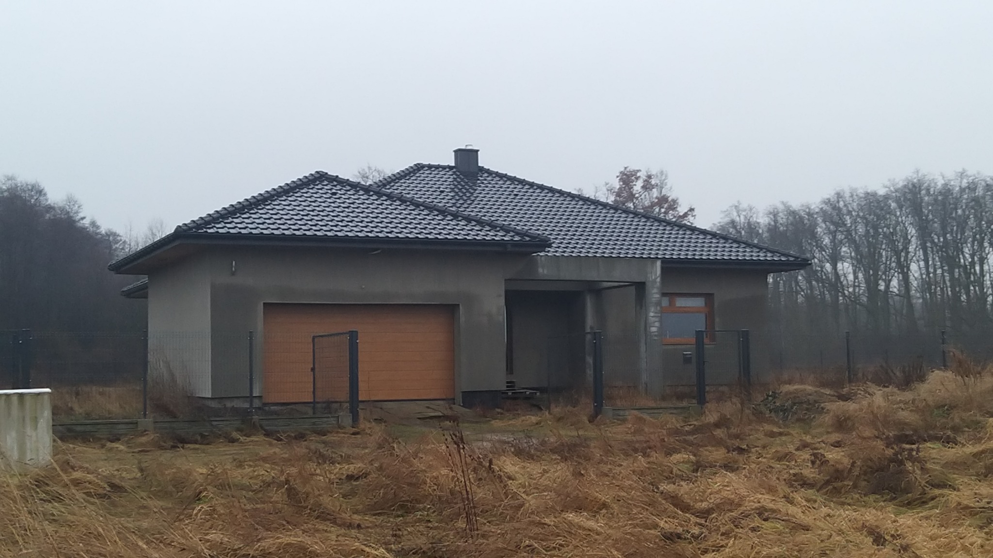 Surowy, parterowy dom jednorodzinny z garażem w stanie deweloperskim, z brązową bramą garażową i ciemną dachówką, otoczony wysoką trawą i ogrodzeniem z siatki, sfotografowany w pochmurny dzień.