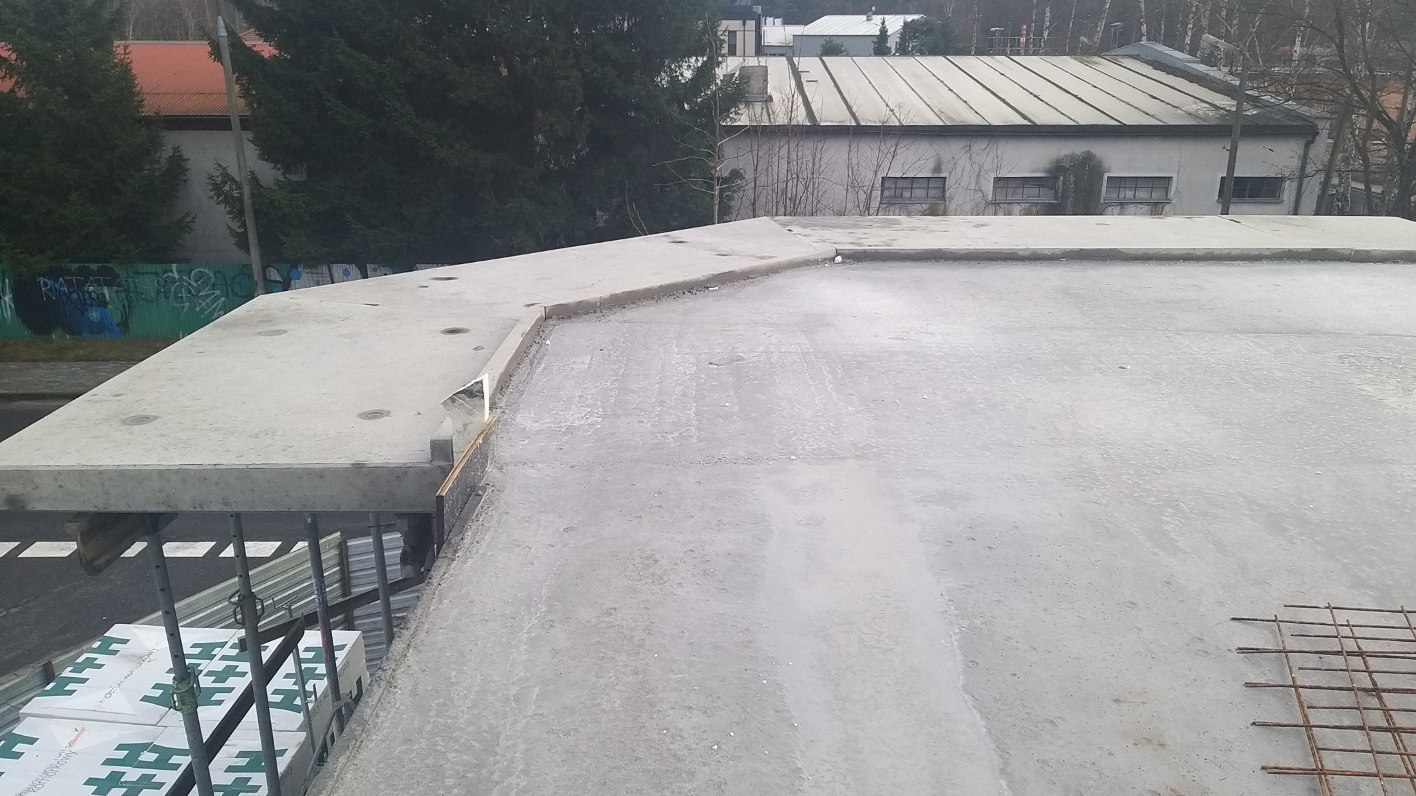 Surowy betonowy strop budynku w trakcie budowy, widoczne podpory szalunkowe i fragment zbrojenia, w tle budynek gospodarczy i drzewa.