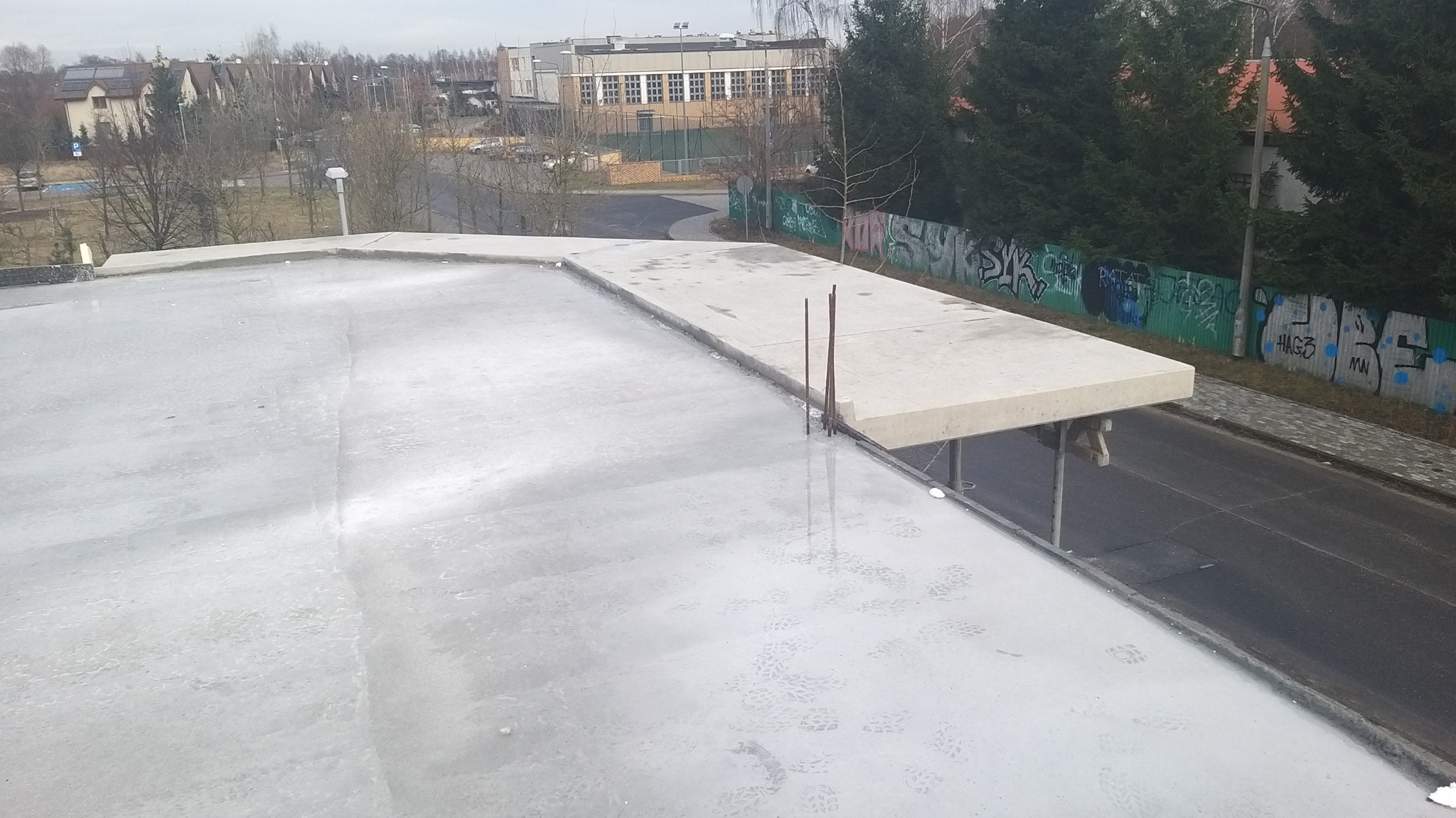 Świeżo wylana, wilgotna płyta betonowa z wystającymi prętami zbrojeniowymi na tle miejskiego krajobrazu z budynkami i drzewami.