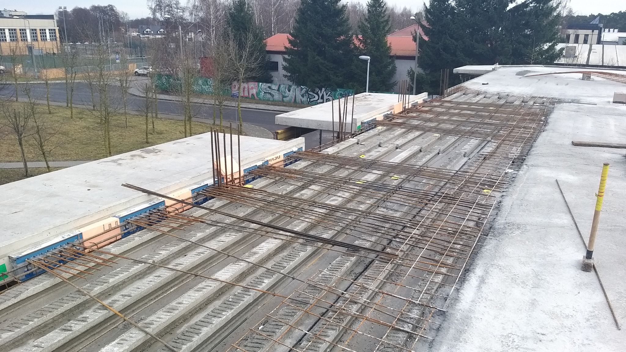 Konstrukcja stropu żelbetowego w trakcie budowy, widoczne prefabrykowane elementy i siatka zbrojeniowa, w tle budynek szkolny i drzewa.