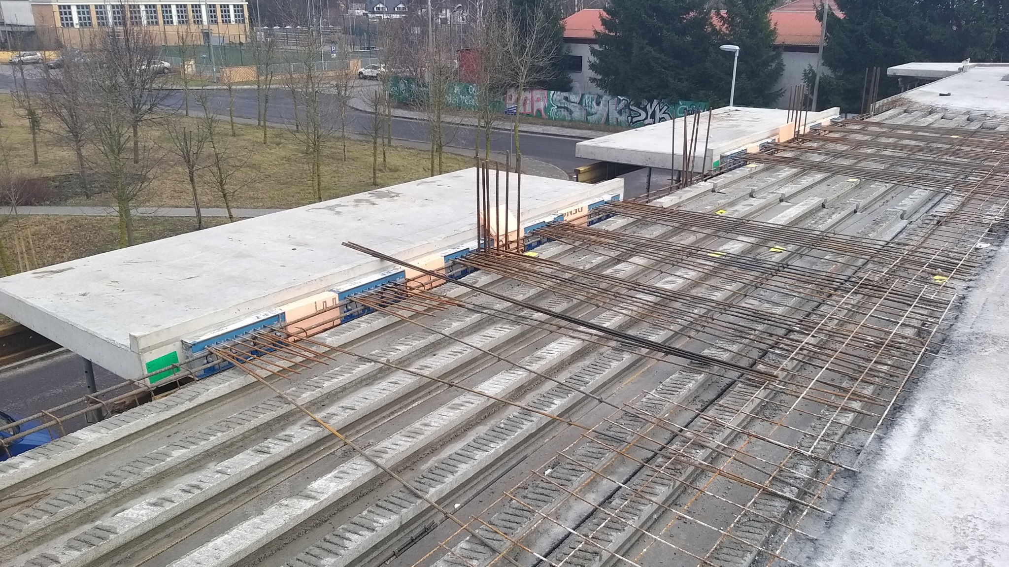 Konstrukcja stropu żelbetowego z widocznym zbrojeniem stalowym i prefabrykatami betonowymi, w tle budynki i drzewa.