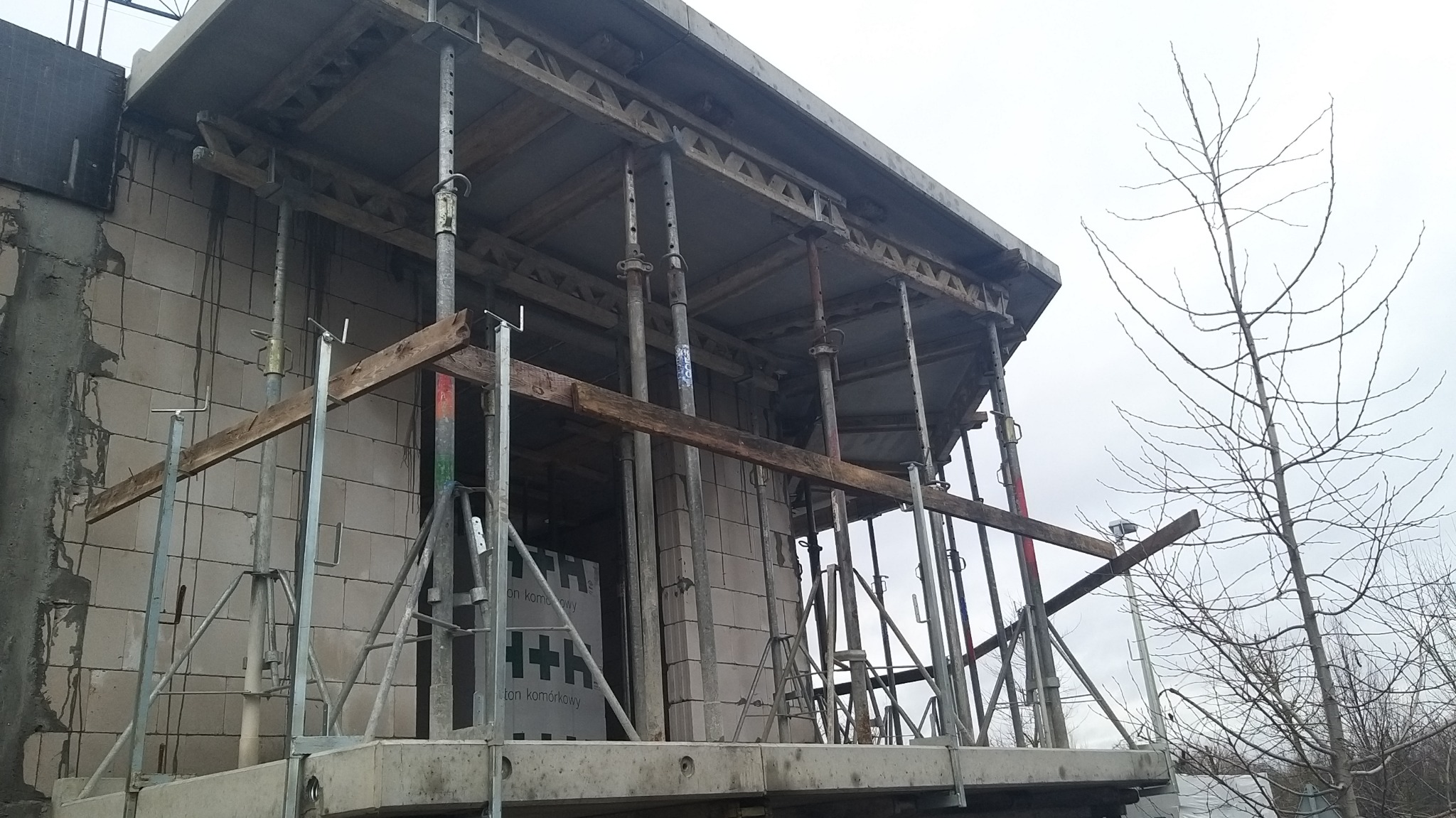 Wznoszenie ścian z betonu komórkowego z użyciem stempli budowlanych i drewnianych belek, widoczna ściana szczytowa budynku w stanie surowym otwartym, na zewnątrz widoczne gołe drzewo.