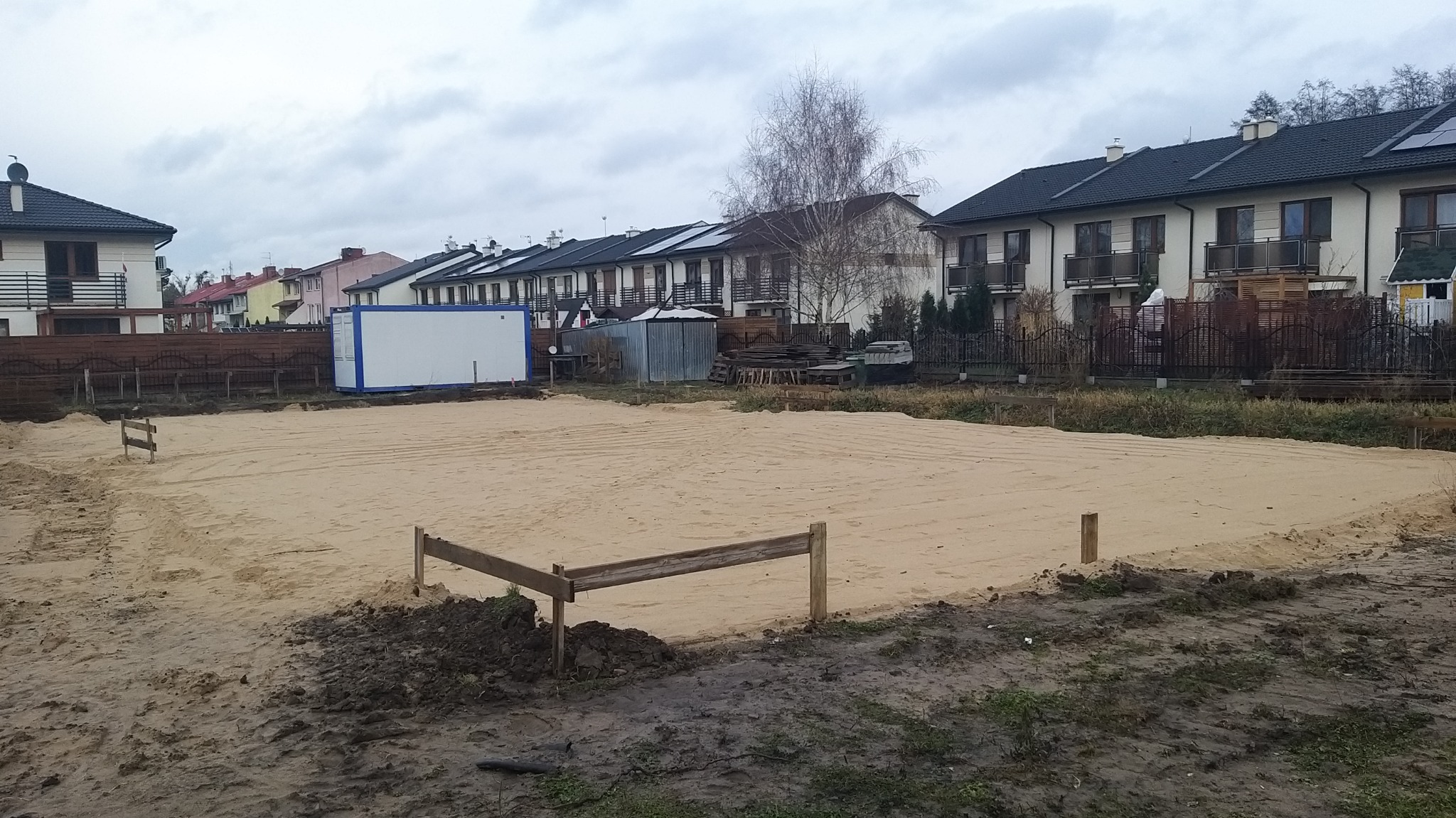 Przygotowany teren pod budowę domu jednorodzinnego z widocznymi drewnianymi słupkami wyznaczającymi obrys fundamentów, w tle zabudowa szeregowa i kontener budowlany.