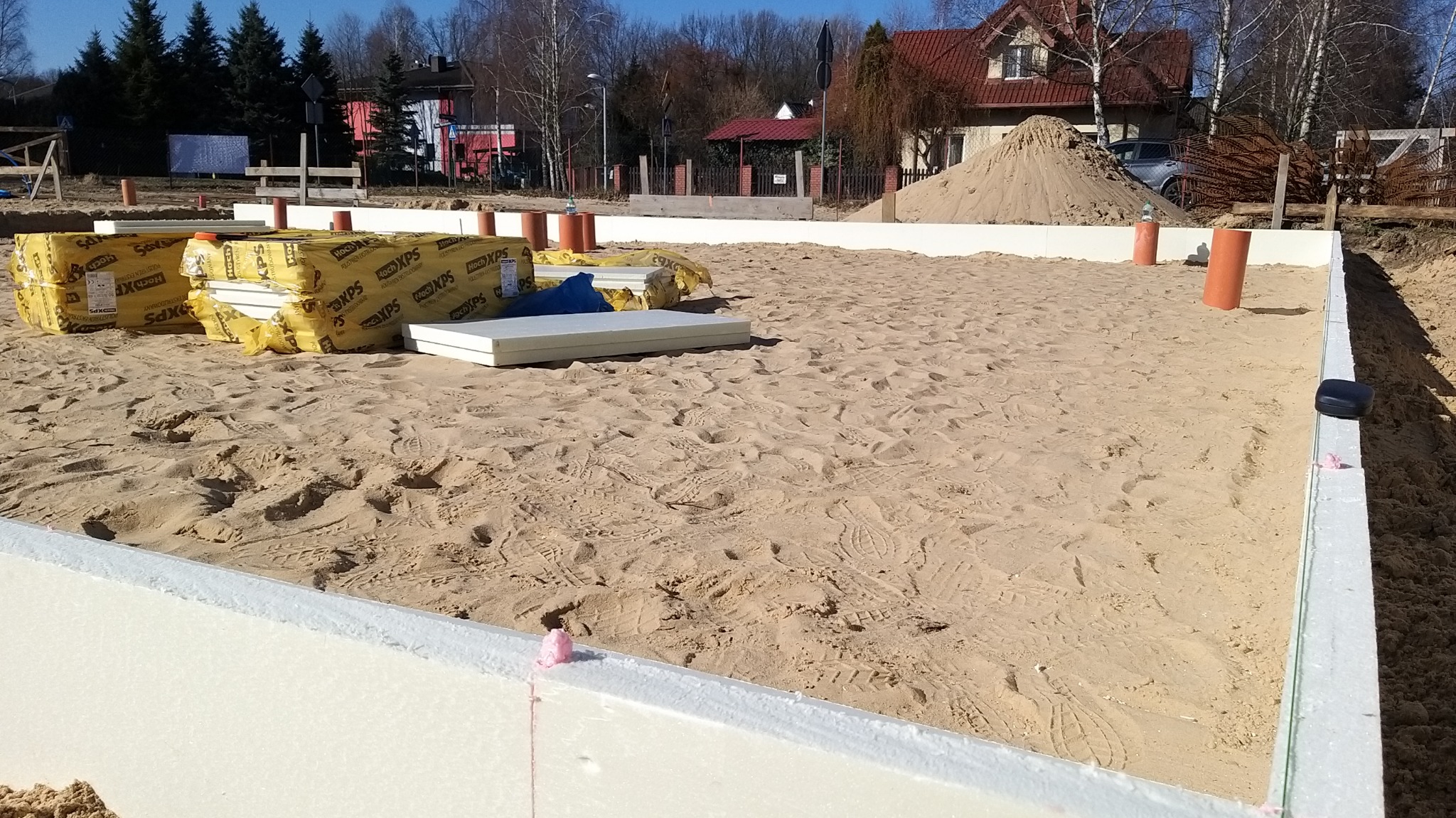 Przygotowany teren pod budowę domu z widocznymi fundamentami w szalunkach z izolacją termiczną, na piasku leżą zapakowane płyty izolacyjne Hoch XPS.