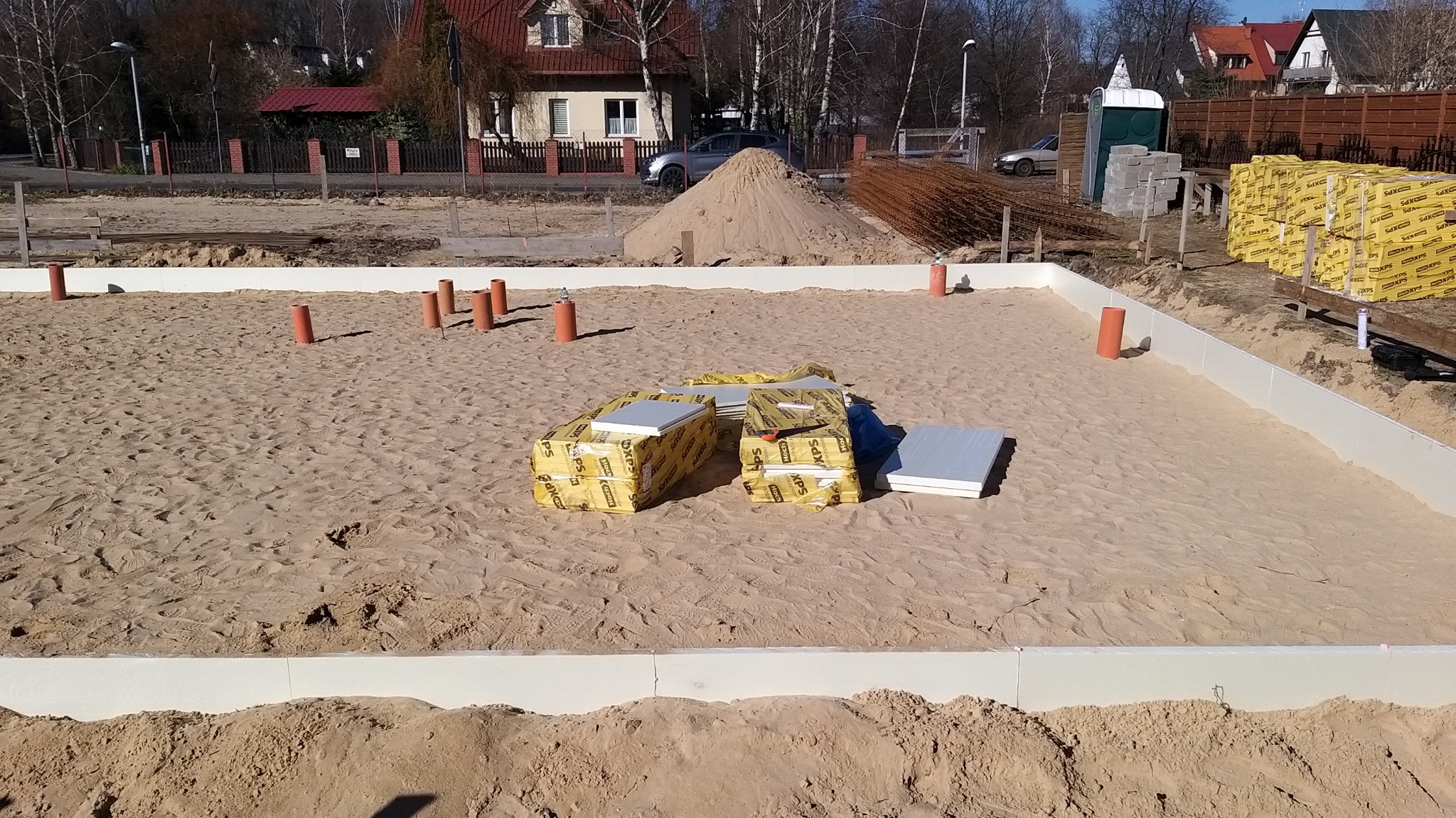 Przygotowany wykop pod fundamenty z widoczną izolacją pionową, rurami instalacyjnymi i stosem paczek izolacyjnych XPS na piasku.
