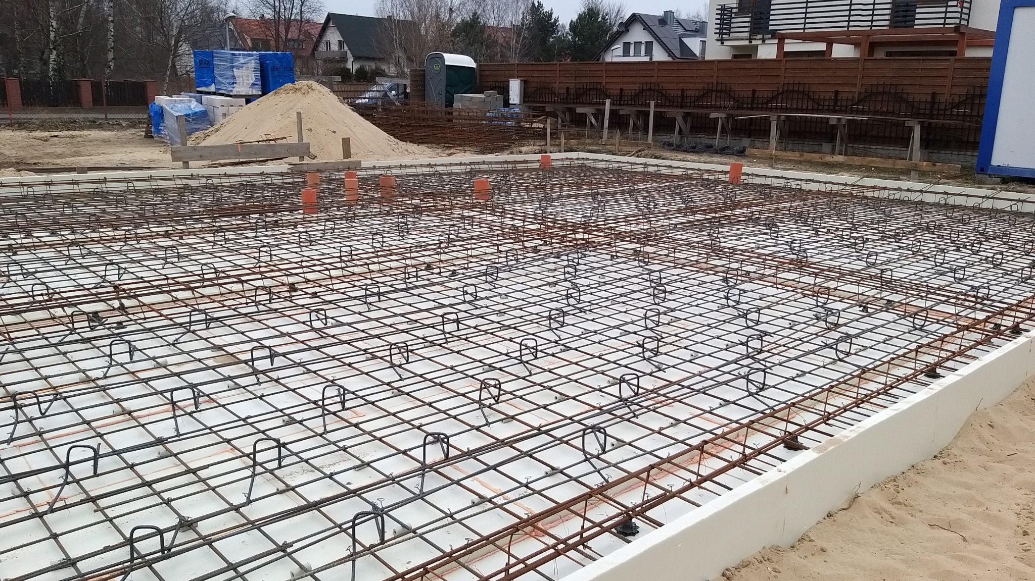 Zbrojenie fundamentu z widoczną siatką stalową, dystansami i wystającymi rurami kanalizacyjnymi na białej izolacji termicznej, w tle sterta piasku, materiały budowlane i zabudowa jednorodzinna.