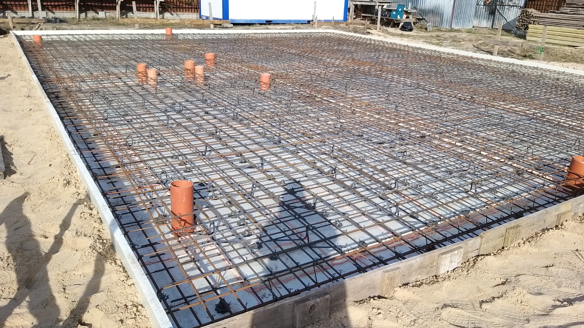 Siatka zbrojeniowa ułożona na fundamencie z widocznymi pionowymi rurami kanalizacyjnymi, otoczona szalunkiem i izolacją, przygotowana do zalania betonem.