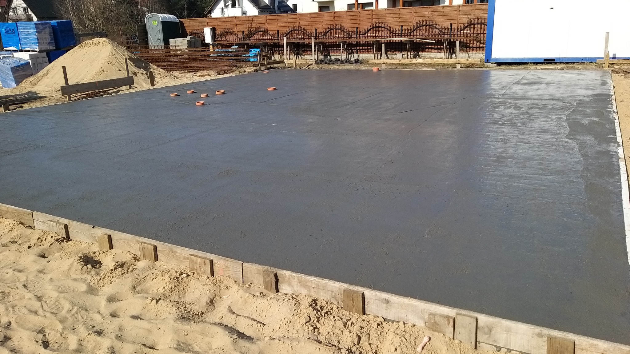Świeżo wylana, gładka płyta fundamentowa z widocznymi deskami szalunkowymi, z tyłu materiały budowlane i przenośna toaleta.