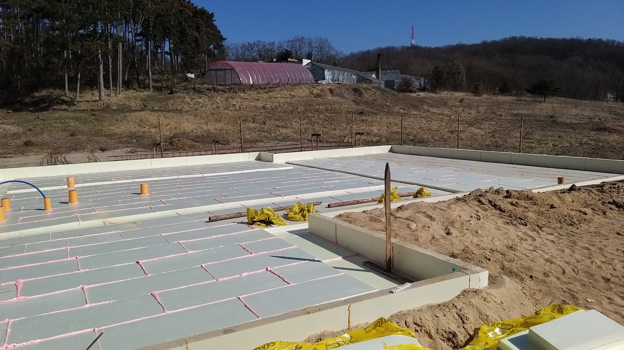 Przygotowanie fundamentów z izolacją termiczną, widoczne płyty styropianowe z różowym uszczelnieniem, rury instalacyjne i szalunki obwodowe, w tle krajobraz z drzewami i budynkami