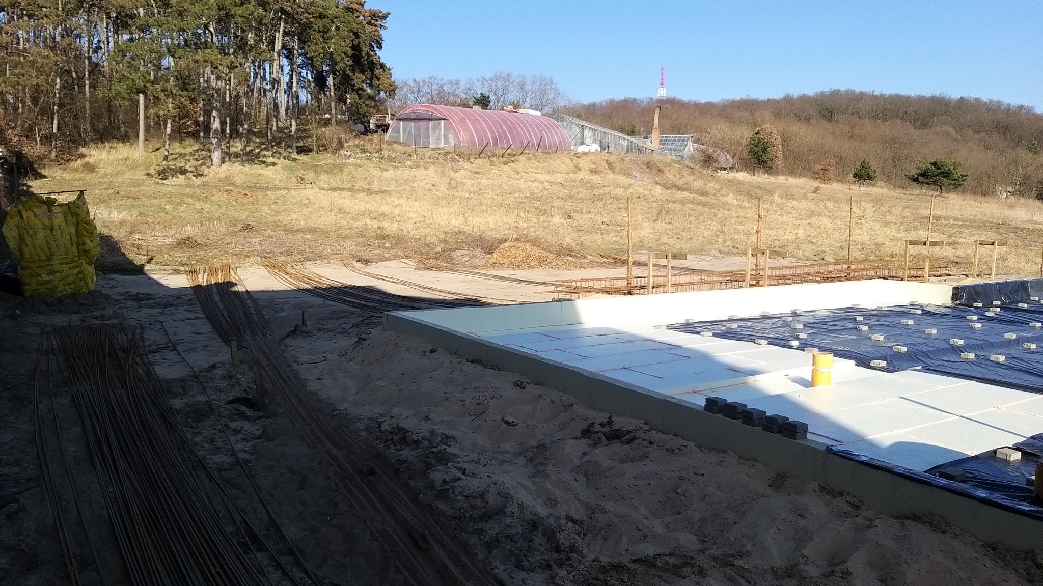 Przygotowany teren pod budowę domu: izolacja fundamentów z użyciem styropianu, folii i obciążników, w tle widoczna konstrukcja szklarni i wieża telekomunikacyjna.