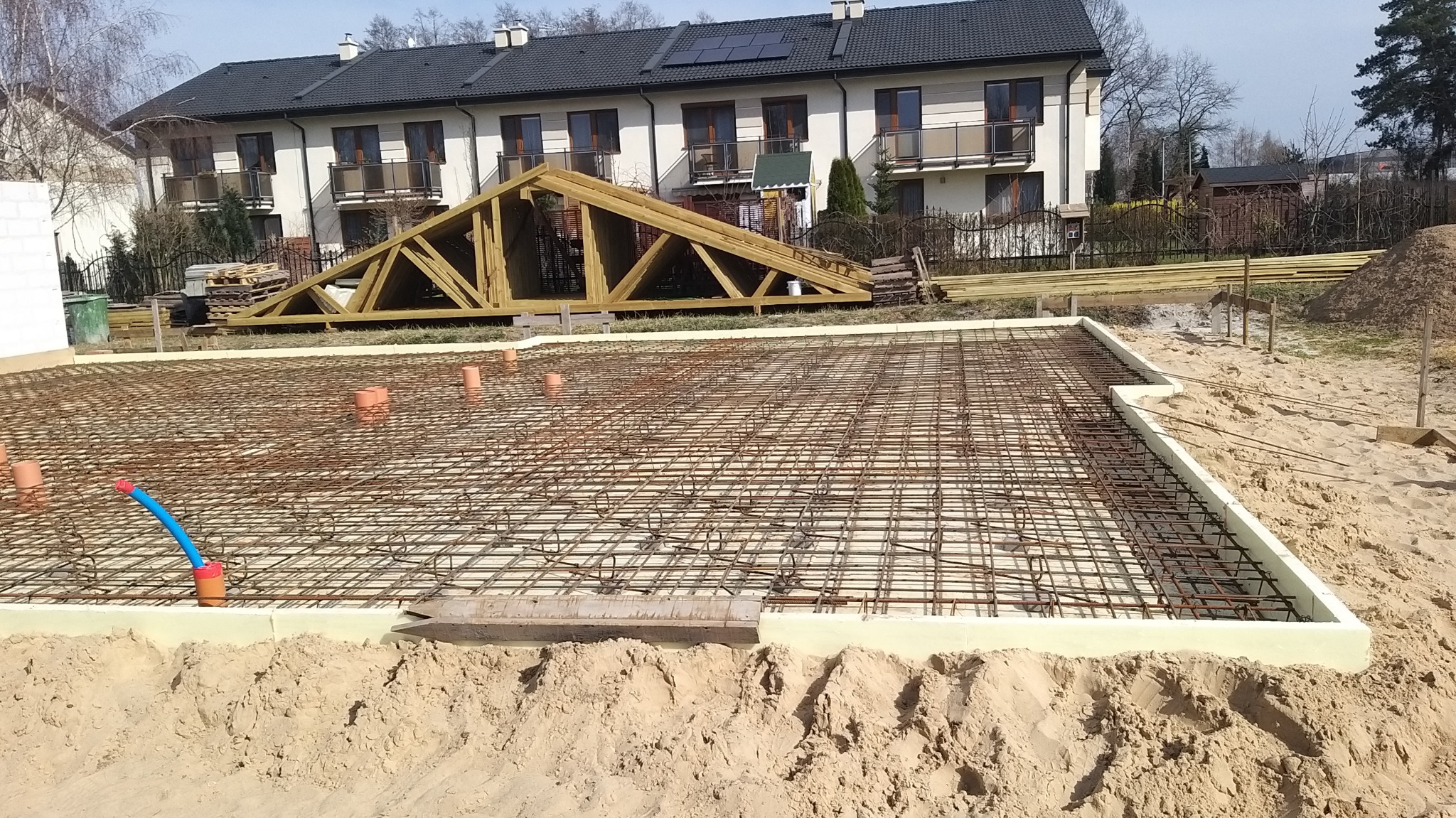 Przygotowany zbrojony fundament z izolacją, widoczne wystające rury instalacyjne, w tle konstrukcja więźby dachowej i budynek mieszkalny.