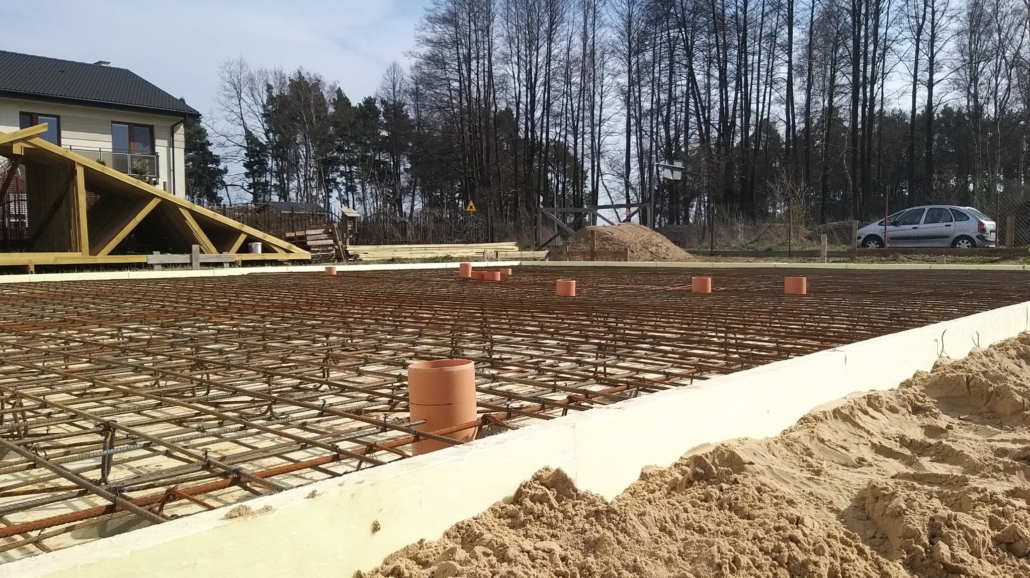 Przygotowany zbrojony fundament pod budowę domu jednorodzinnego, widoczne szalunki, siatka zbrojeniowa, rury kanalizacyjne oraz nasyp piasku.