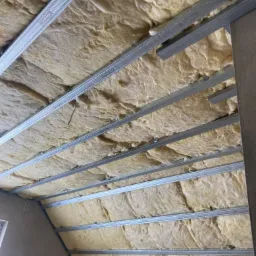 Poddasze w trakcie ocieplania wełną mineralną, widoczne metalowe profile mocujące, fragment okna po lewej stronie.