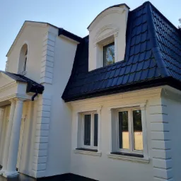 Biała elewacja domu z czarną dachówką, widoczne kolumny przy wejściu i ozdobne obramowania okien.