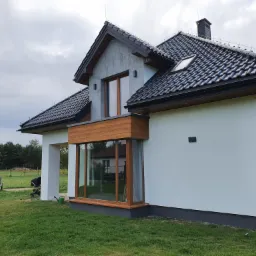 Nowoczesny dom jednorodzinny z białą elewacją i drewnianą stolarką okienną, widok z boku na zadbany trawnik z meblami ogrodowymi i wózkiem dziecięcym.