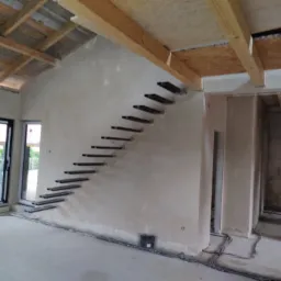 Surowy tynk maszynowy na ścianie wewnętrznej w trakcie budowy, z zamontowanymi schodami wspornikowymi i widoczną instalacją elektryczną przy podłodze.