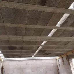 Wnętrze budynku w trakcie budowy: sufit z betonowych bloków wsparty na drewnianych belkach, ściany wzniesione z białych bloczków.