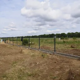 Nowo postawione ogrodzenie panelowe z podmurówką betonową na działce z widokiem na pole i las w tle, w pochmurny dzień.