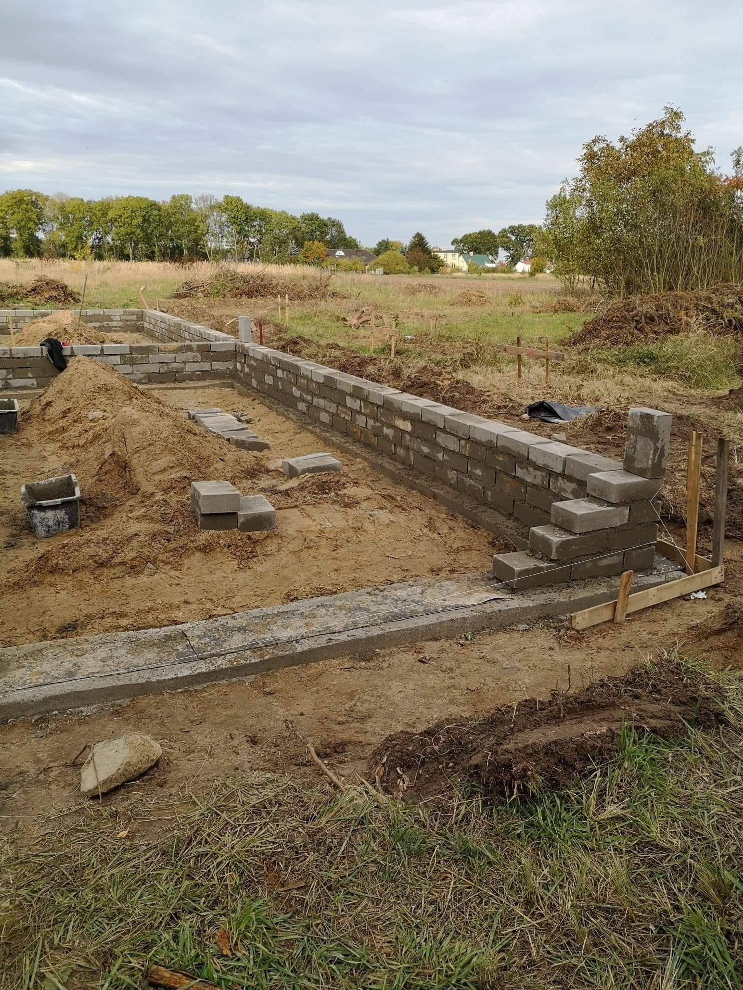 Wznoszenie muru fundamentowego z bloczków betonowych na działce budowlanej, widoczne szalunki i przygotowany teren pod budowę.