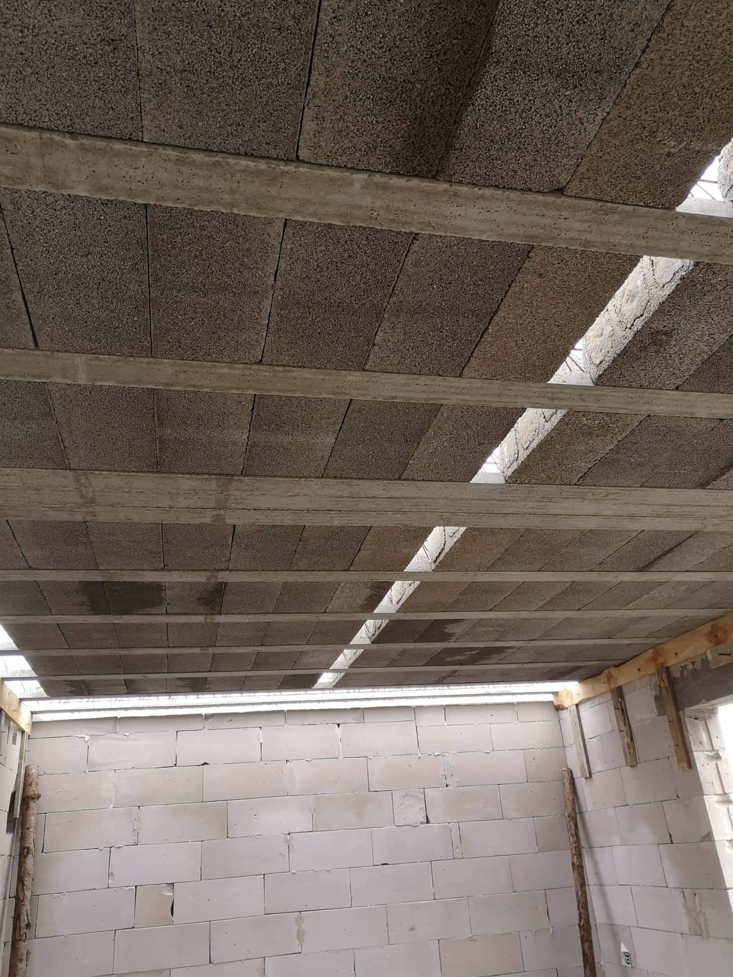 Wnętrze budynku w trakcie budowy: sufit z betonowych bloków wsparty na drewnianych belkach, ściany wzniesione z białych bloczków.