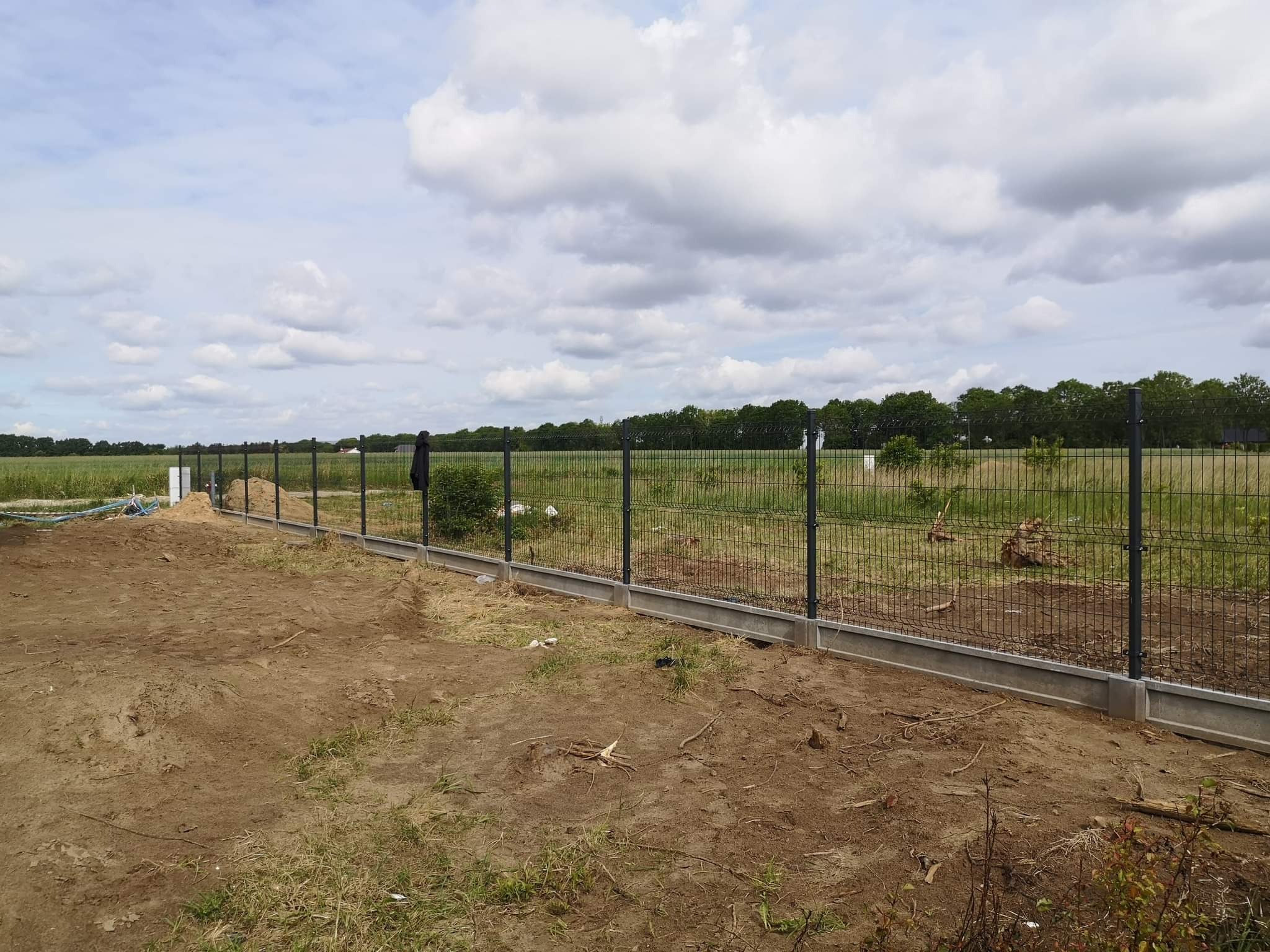 Nowo postawione ogrodzenie panelowe z podmurówką betonową na działce z widokiem na pole i las w tle, w pochmurny dzień.