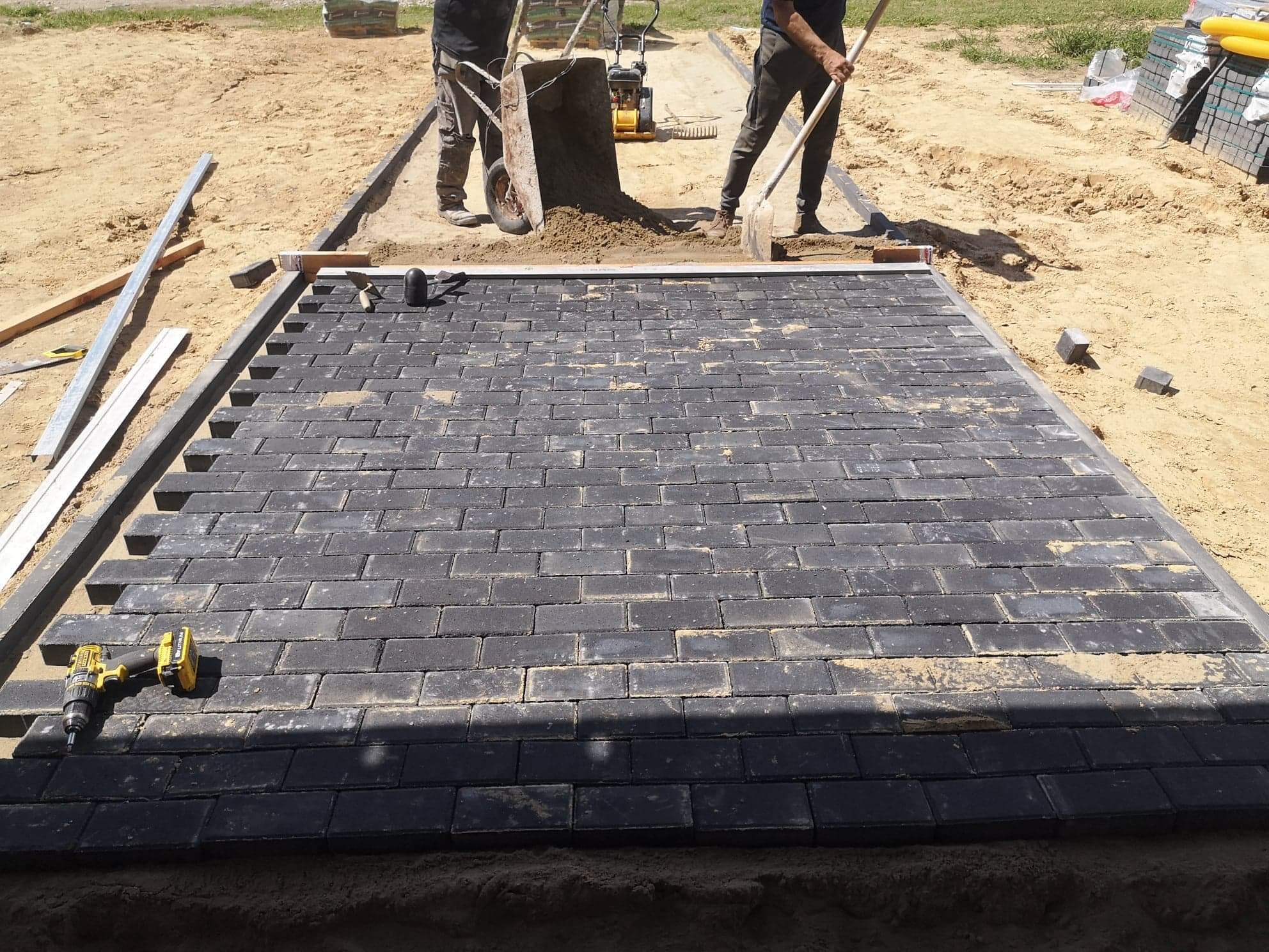 Częściowo ułożona, prostokątna nawierzchnia z ciemnej kostki brukowej, z widocznymi narzędziami (wiertarka, młotek) i pracownikami w tle, przygotowującymi podłoże.