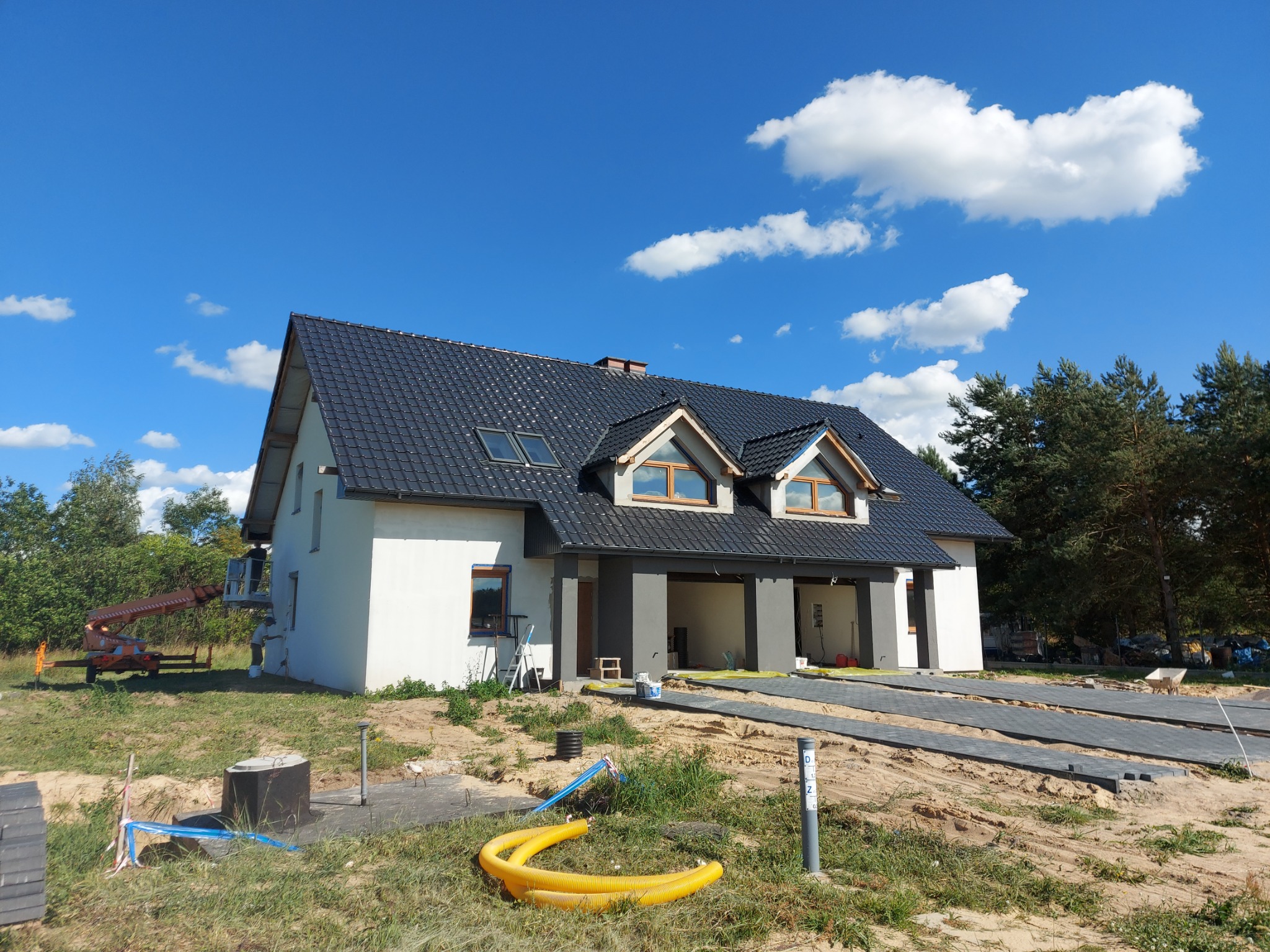 Nowo wybudowany dom jednorodzinny z ciemną dachówką, widoczna elewacja w trakcie wykończenia z użyciem podnośnika koszowego, przygotowane podłoże pod przyszły podjazd z kostki brukowej.