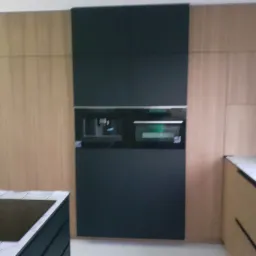 Nowoczesna zabudowa kuchenna z forniru w odcieniach beżu i czarnym panelem z wbudowanymi sprzętami AGD, widoczna część blatu z szarego kamienia.