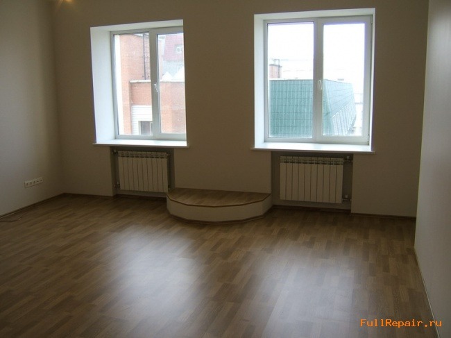 Pusta przestrzeń w pokoju z dwoma oknami, parkietem ułożonym w deski i półokrągłym podestem między grzejnikami.