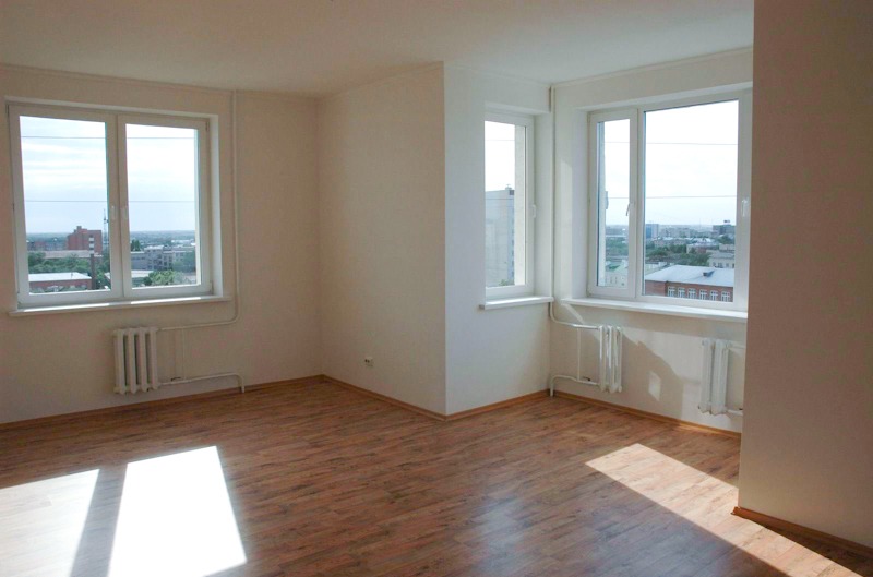 Pusty pokój po remoncie z panelami podłogowymi w odcieniach brązu, białymi ścianami i grzejnikami pod dwoma oknami z widokiem na miasto.