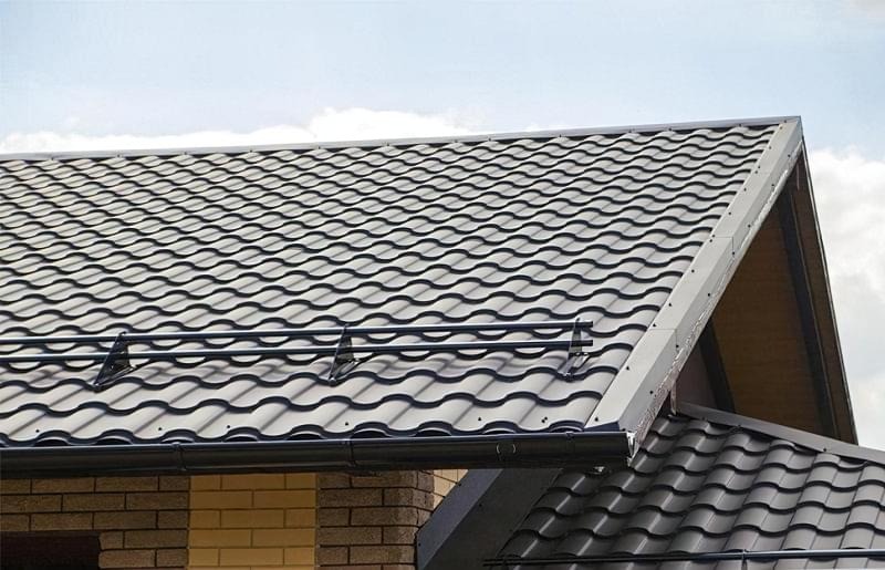 Nowoczesne, metalowe pokrycie dachowe w kolorze grafitowym z systemem rynnowym, widoczne z bliska pod kątem, z fragmentem murowanej ściany poniżej.