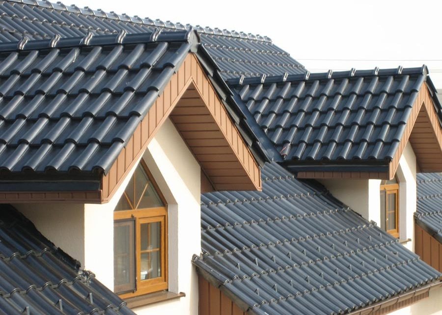 Fragment dachu z ciemną dachówką ceramiczną i drewnianymi elementami wykończeniowymi, widoczne okna w lukarnach, jasna elewacja budynku.