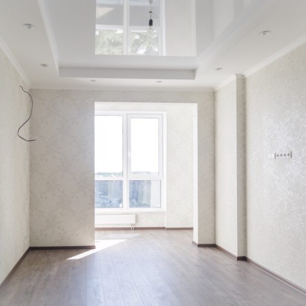 Pusty pokój po odświeżeniu, z ciemnymi panelami podłogowymi, jasnymi ścianami z delikatnym wzorem, białym sufitem podwieszanym z oświetleniem punktowym, widocznymi gniazdkami elektrycznymi...