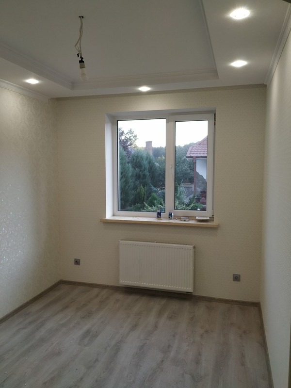 Pokój po odświeżeniu z panelami podłogowymi, grzejnikiem pod oknem i ozdobną listwą sufitową, widoczna instalacja elektryczna w suficie przed montażem lampy.