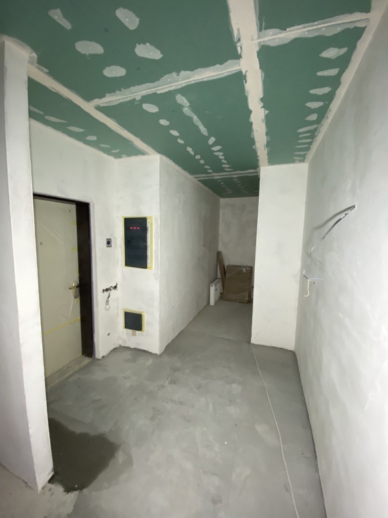 Wnętrze w trakcie prac wykończeniowych: sufit z płyt gipsowo-kartonowych, świeżo zagruntowane ściany, wylewka podłogowa, widoczna instalacja elektryczna, drzwi wejściowe z ciemną ościeżnicą po...