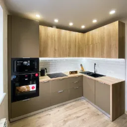 Nowoczesna kuchnia narożna z frontami w kolorze taupe i drewnopodobnymi szafkami górnymi, oświetleniem podszafkowym LED i sprzętem AGD w zabudowie, widok na czarny zlew i płytę indukcyjną.