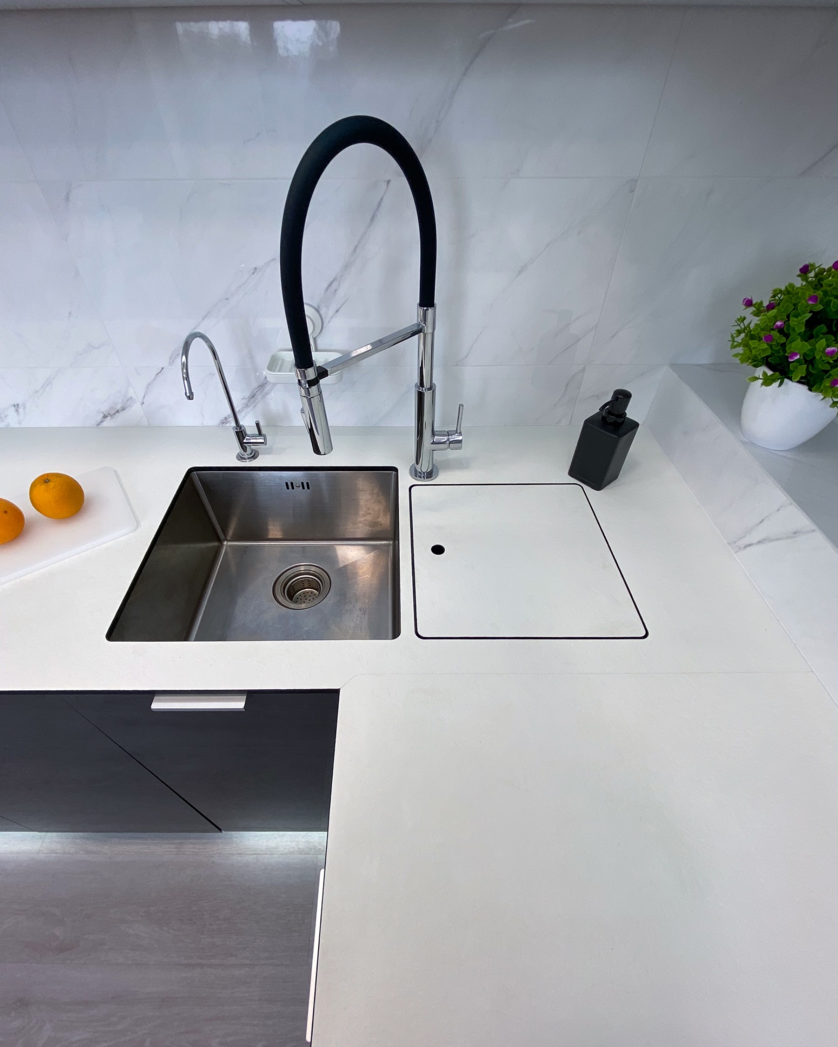 Nowoczesny zlew kuchenny ze stali nierdzewnej z czarną, elastyczną wylewką, wbudowany w biały blat z marmurowym wzorem, obok deska do krojenia z pomarańczami i dozownik do mydła.