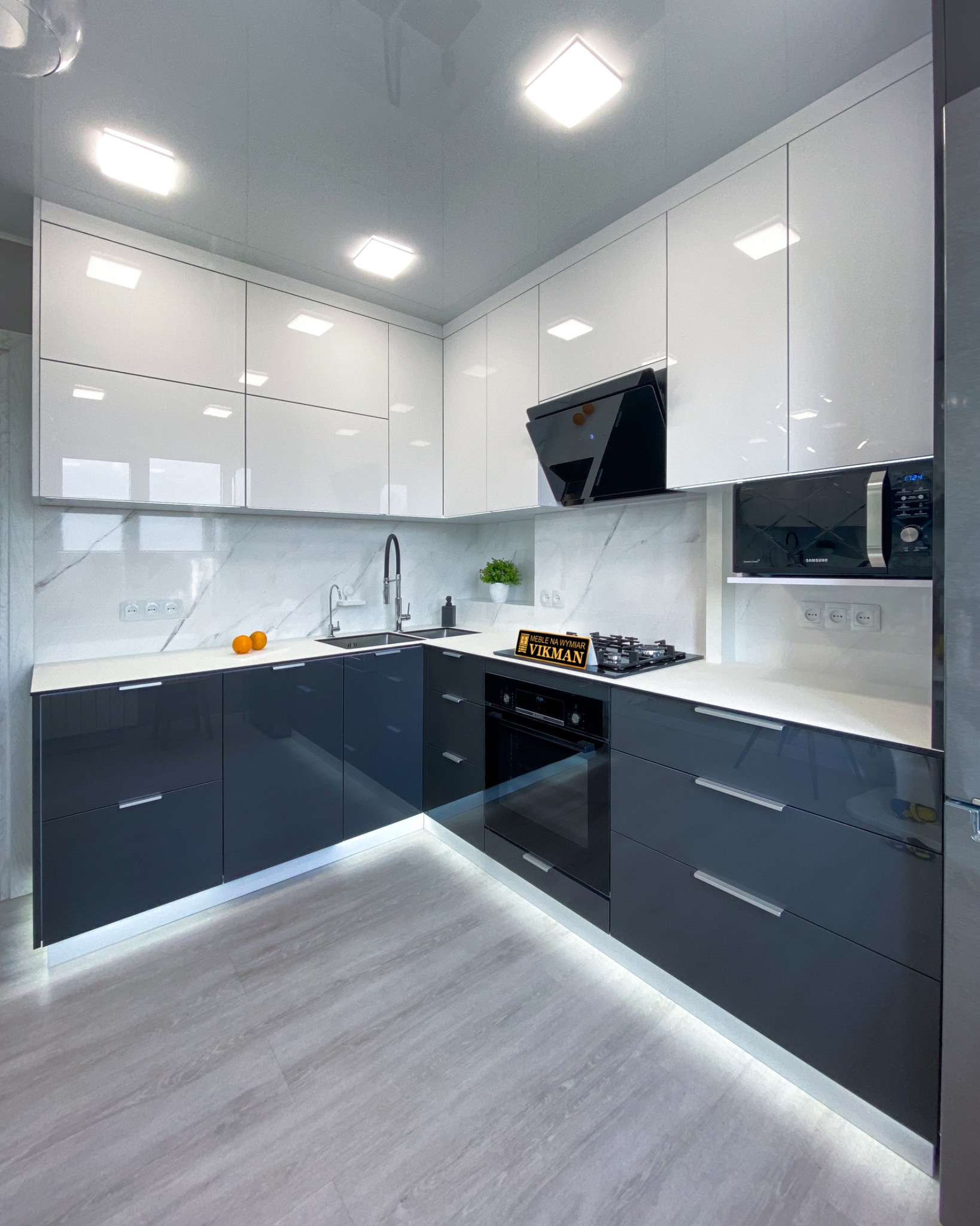 Nowoczesna kuchnia w odcieniach bieli i grafitu z połyskiem, podświetleniem LED u dołu szafek, marmurowym backsplashiem i blatem, z czarnym okapem i piekarnikiem w zabudowie, oraz drobną dekoracją...