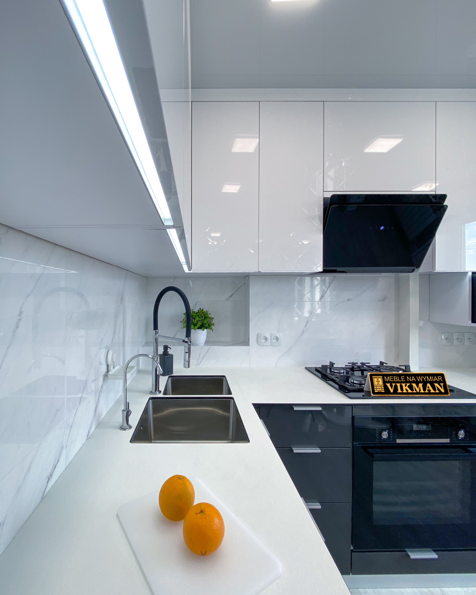 Nowoczesna kuchnia z białymi, lakierowanymi szafkami górnymi i czarnymi dolnymi, marmurowy backsplash, stalowe zlewozmywaki, czarna bateria i płyta grzewcza. Na blacie deska z dwoma pomarańczami.