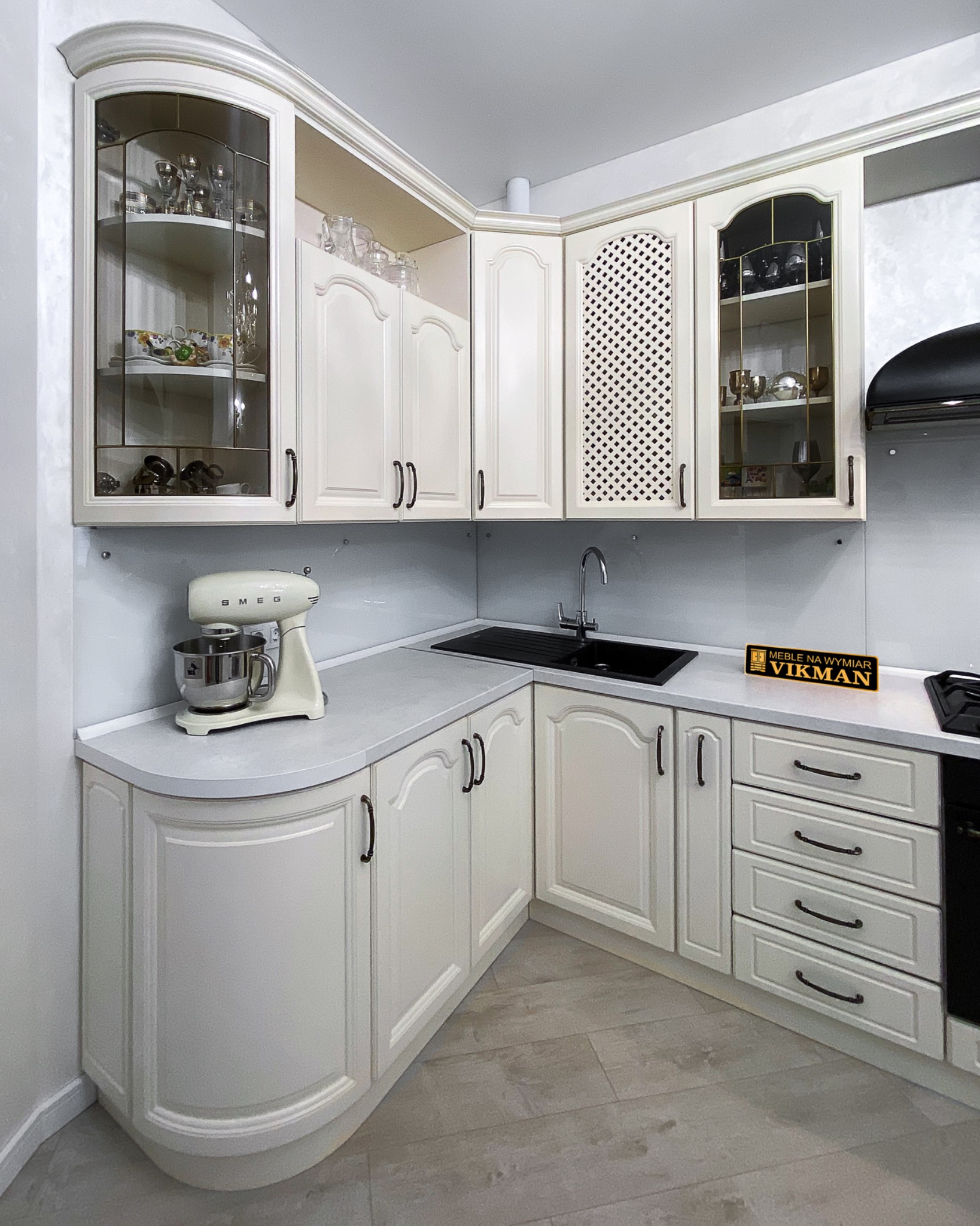 Jasna, klasyczna zabudowa kuchenna z frezowanymi frontami, witrynami ze szprosami i ażurowymi wstawkami, uzupełniona o czarny zlew, baterię i sprzęt AGD SMEG, z widocznym logo firmy na blacie.