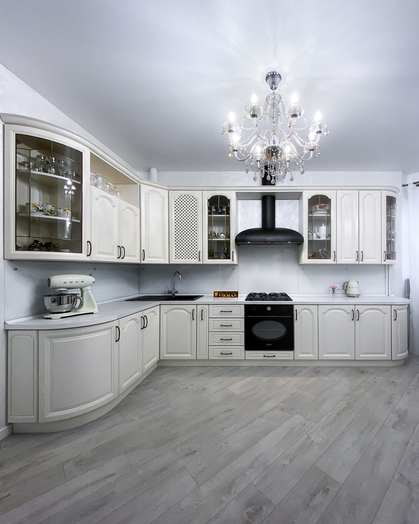 Elegancka, jasna kuchnia z białymi frontami, czarnym zlewem i piekarnikiem Smeg, oświetlona kryształowym żyrandolem, z drewnianą podłogą ułożoną w jodełkę.