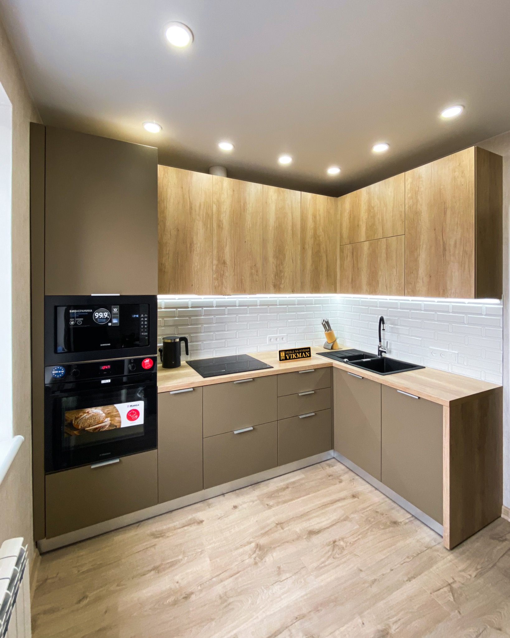 Nowoczesna kuchnia narożna z frontami w kolorze taupe i drewnopodobnymi szafkami górnymi, oświetleniem podszafkowym LED i sprzętem AGD w zabudowie, widok na czarny zlew i płytę indukcyjną.