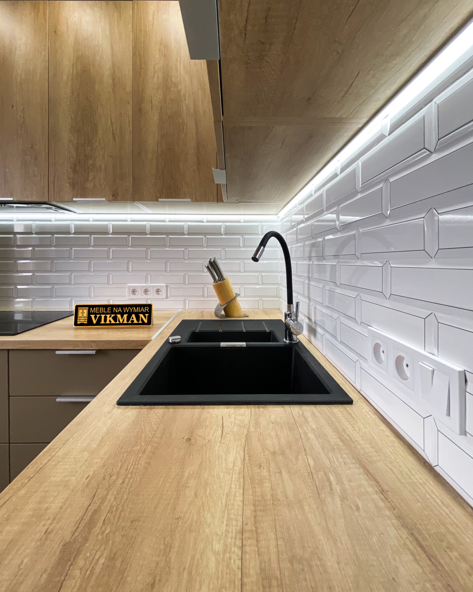 Czarny, dwukomorowy zlew wpuszczony w drewniany blat, czarna bateria, białe płytki na ścianie i oświetlenie LED podszafkowe w kuchni w Gdyni.