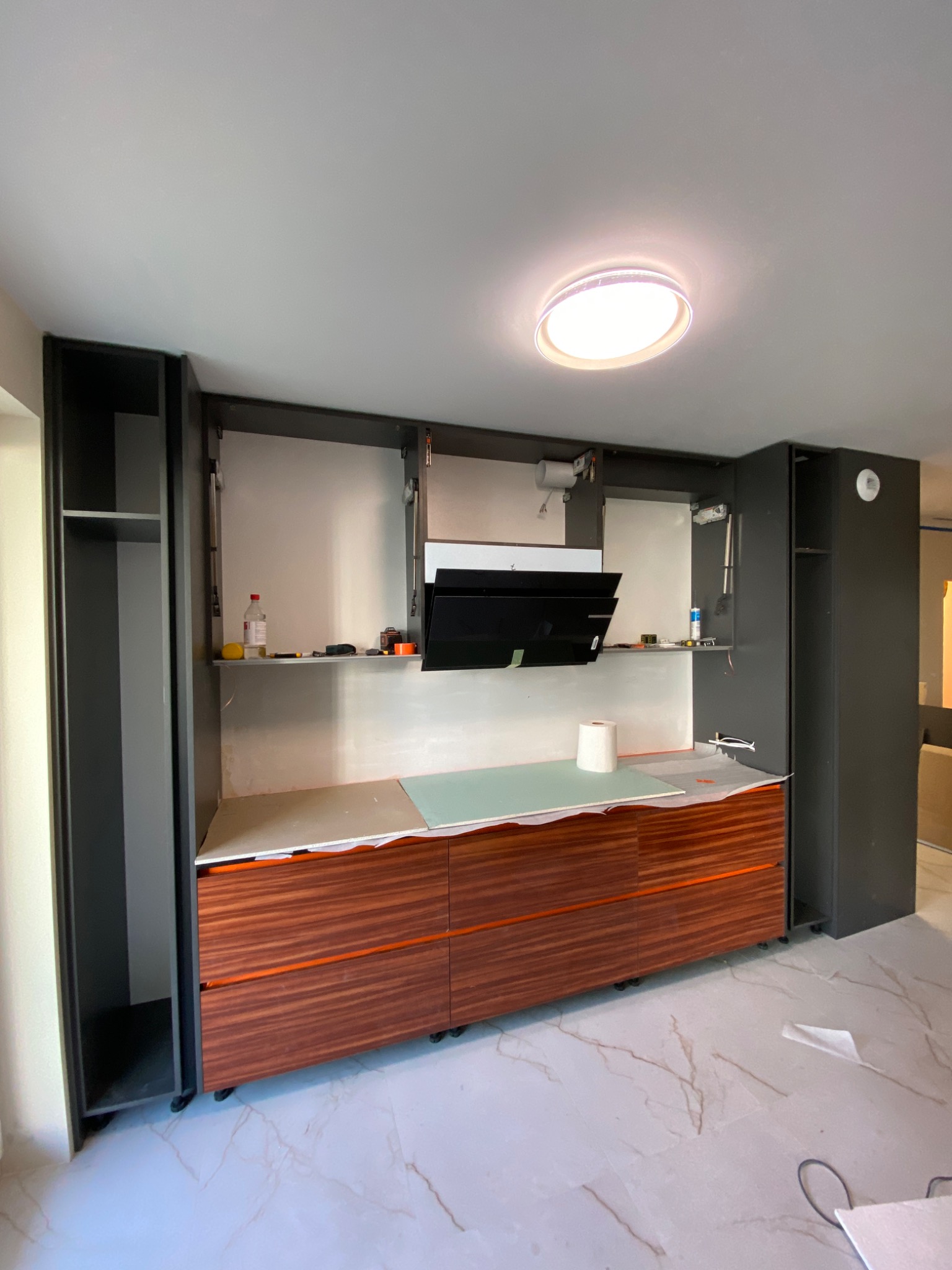 Montaż szafek kuchennych: widoczne brązowe fronty dolnych szafek z pomarańczowymi wstawkami, szare szafki górne bez frontów, czarny okap, biały blat zabezpieczony folią, marmurowe płytki...
