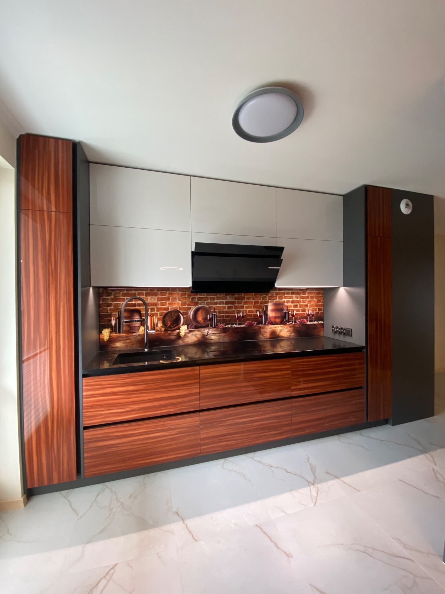 Nowoczesna kuchnia z frontami w kolorze drewna i szarymi elementami, czarny blat i okap, na ścianie nad blatem fototapeta z motywem wina i beczek.