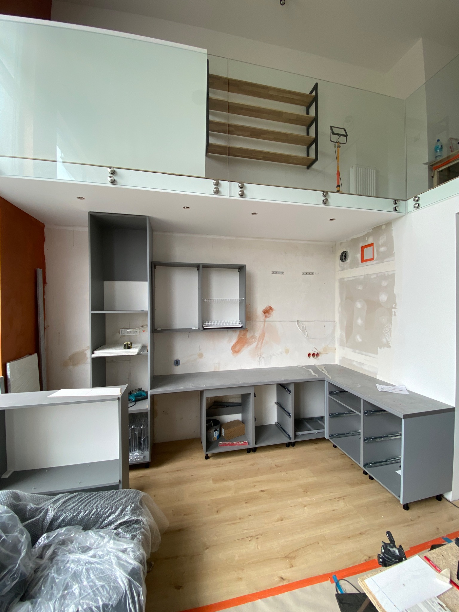 Montaż szarej zabudowy kuchennej w surowym stanie, widoczne niedokończone ściany, elementy szklanej balustrady na antresoli, narzędzia i materiały budowlane na podłodze.