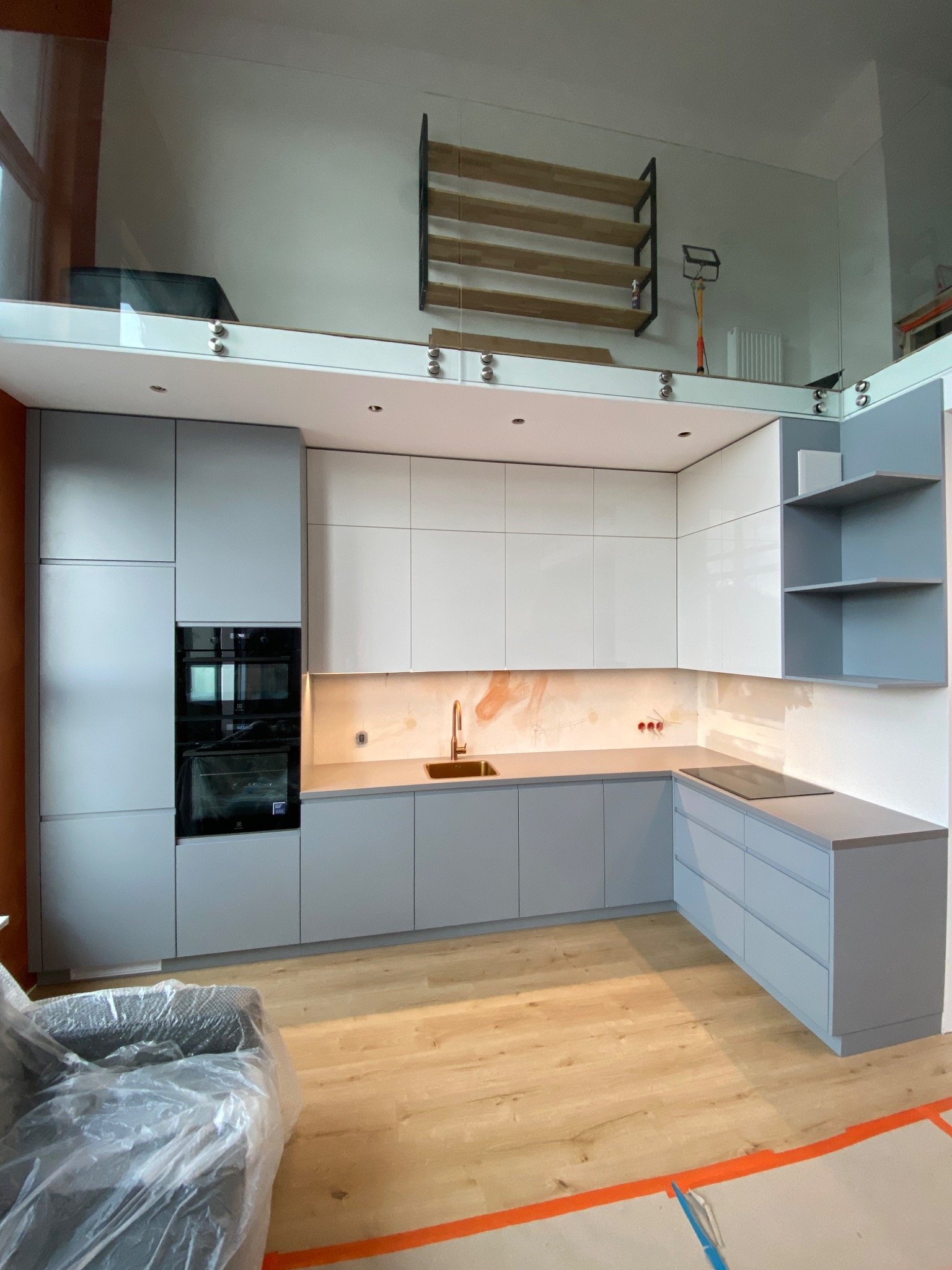 Nowoczesna kuchnia w odcieniach szarości i bieli z drewnianą podłogą i przeszkloną antresolą, widoczne zabudowane sprzęty AGD i elementy wykończeniowe, meble zakryte folią ochronną.