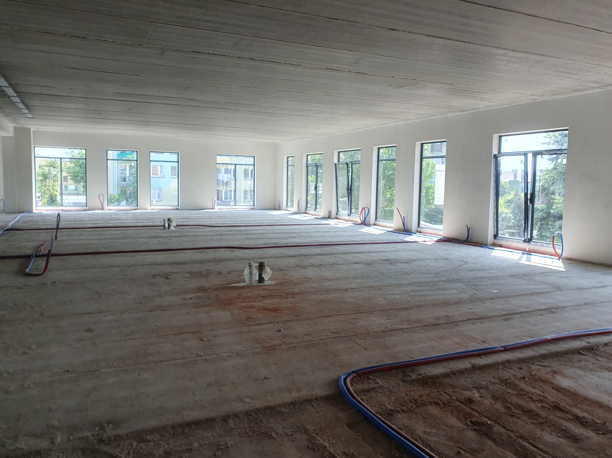Wnętrze w stanie deweloperskim z instalacją ogrzewania podłogowego. Widoczne rury grzewcze na surowej posadzce i duże okna z widokiem na zieleń.
